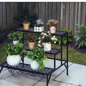 May include: A black, three-tiered plant stand with decorative scrollwork. The stand holds various potted plants and floral arrangements in white, pink, and beige pots. The stand is made of metal and is placed on a stone patio.