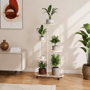 Op de afbeelding: Een hoge, gebroken witte metalen plantenstandaard met zes ronde planken op verschillende hoogtes. De structuur wordt ondersteund door een dun, gebogen frame en staat op kleine zwarte wieltjes, gevuld met diverse groene planten in potten.