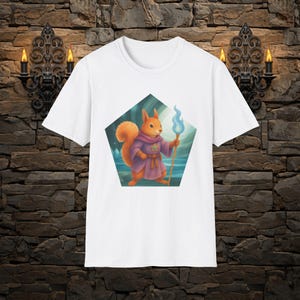 May include: A white t-shirt featuring a pentagonal graphic of an orange squirrel dressed in a purple robe with a gold star emblem. The squirrel holds a wooden staff topped with a glowing blue flame against a backdrop of a misty forest landscape.
