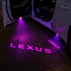 Puede incluir: Un vehículo Lexus blanco con puertas abiertas que cuenta con luces LED púrpuras que proyectan la palabra LEXUS en el suelo. El tablero, el volante y los pedales son visibles a través de la puerta del conductor abierta durante la noche.