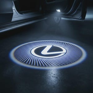 Puede incluir: Un proyector de luz de cortesía para puerta de coche que proyecta un logotipo de Lexus blanco brillante en el suelo. La proyección circular presenta un emblema L central rodeado por un patrón radial detallado de puntos desde la puerta abierta.