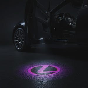 Puede incluir: Un vehículo de color oscuro con la puerta del conductor abierta, que cuenta con una luz de cortesía que proyecta un logotipo de Lexus circular, brillante, de color púrpura y rosa, en el suelo. El coche tiene llantas plateadas y un volante visible.