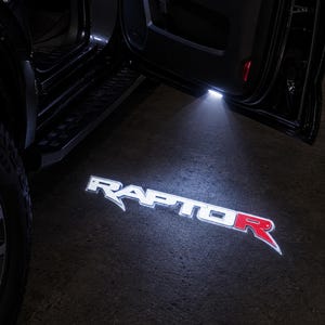 Può includere: Un proiettore di luce per portiera di veicolo che proietta un logo Raptor bianco e rosso brillante a terra. La luce emana dalla parte inferiore di una portiera aperta di un'auto nera, illuminando la superficie scura con il testo grafico.
