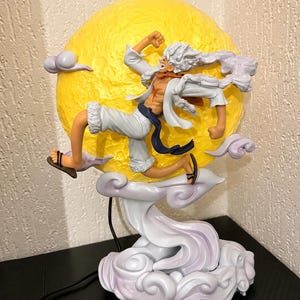 Könnte beinhalten: Eine dynamische Anime-Figurenstatue mit stacheligen weißen Haaren, einem offenen weißen Hemd, weißen Shorts und einer dunklen Schärpe, die vor einem großen, texturierten gelben Mond springt. Der Sockel besteht aus wirbelnden hellvioletten Wolken.