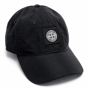 May include: A black baseball cap featuring a square patch on the front panel. The patch displays a white circular compass logo with the text Stone Island. The cap has a curved brim, stitched panels, and small ventilation eyelets on the crown.