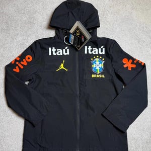 Puede incluir: Una chaqueta negra con capucha que presenta el logotipo blanco de Itaú, un logotipo amarillo de Jumpman y el escudo de la selección nacional de fútbol de Brasil en el pecho. Las mangas muestran gráficos naranjas de Vivo y figuras humanas.