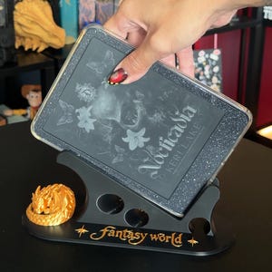 Pode incluir: Uma capa preta com glitter para e-reader com o título do livro Nocticadia de Keri Lake, exibida num suporte preto com uma figura de dragão dourado e o texto Fantasy world impresso a dourado na base, com um design estético escuro.