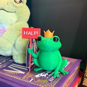 Puede incluir: Una pequeña figura de rana verde con grandes ojos negros y una corona dorada en la cabeza. La rana sostiene un letrero rectangular rojo en un palo delgado que presenta el texto blanco HALP! en letras mayúsculas, impreso en una fuente sans-serif.