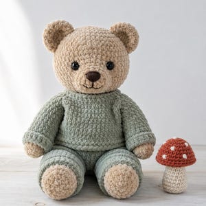 Op de afbeelding: Een handgemaakte gehaakte teddybeer in bruin en saliegroen, met een bijpassende paddenstoel. De beer draagt een saliegroene trui en broek. De paddenstoel heeft een rode hoed met witte stippen en een crème steel.