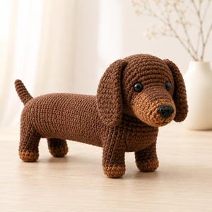 May include: A crocheted dachshund dog in shades of brown. The dog has a long body, short legs, and floppy ears. The eyes are black, and the nose is a darker brown. The dog is standing on a light-coloured surface.