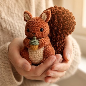 May include: A handmade crochet squirrel in shades of brown, holding a small acorn. The squirrel has a fluffy tail and is held in cupped hands. The acorn is green and yellow. A cozy, handcrafted item.
