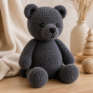 Op de afbeelding: Een donker antracietgrijze gehaakte teddybeer met een rond hoofd, kleine oortjes en een zwart geborduurde neus. Het knuffeldier heeft een textuur van garen, twee donkere plastic ogen en een zittende houding met ronde ledematen en een romp.