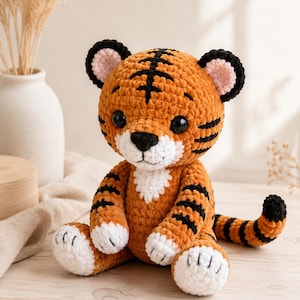 May include: A plush tiger toy made of orange, black, and white yarn. The tiger has a round head with black stripes, black ears with pink centers, and large black eyes. It features a white muzzle, chest patch, and paws with black embroidered details.