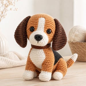 May include: A handmade crochet beagle dog with brown, white, and black details. The dog has large black eyes, floppy brown ears, and a brown collar. The crochet dog is sitting on a light-colored wooden surface.