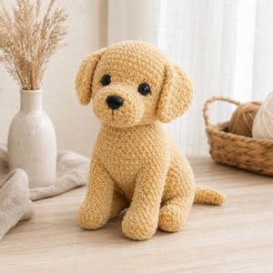 Puede incluir: Un cachorro de perro de color amarillo dorado tejido a ganchillo con ojos y nariz negros, sentado erguido. El animal de peluche hecho a mano tiene orejas caídas y una cola pequeña. El perro está sentado sobre una superficie de madera de color claro.