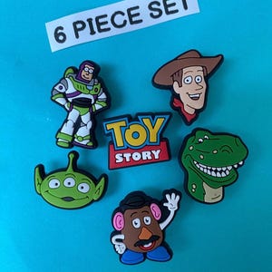 May include: A collection of six vibrant character charms from the animated film Toy Story. The set features Buzz Lightyear, Woody, an alien, Rex, Mr. Potato Head, and the Toy Story logo. The text "6 PIECE SET" is visible.