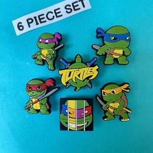Puede incluir: Un conjunto de seis dijes con temática de las Tortugas Ninja. Los dijes presentan a las tortugas en varias poses, cada una con una máscara de diferente color. El conjunto incluye un dije con el logotipo y un dije que dice "6 PIECE SET".