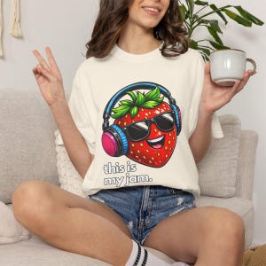 May include: Cream-colored t-shirt featuring a cartoon strawberry wearing sunglasses and headphones. The strawberry has a smiling face and the text "this is my jam" is below the graphic. The person is wearing denim shorts.