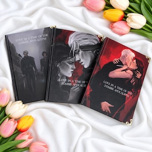 May include: Three hardcover books titled Love in a Time of the Zombie Apocalypse with illustrated covers featuring characters in dark, dramatic scenes. The books have gold-toned metal corner protectors and are arranged on a white fabric surface.