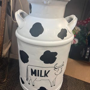 May include: A white metal milk can featuring black cow-print spots and a black line drawing of a cow with the word MILK printed on its side. The container has two side handles and a wide rimmed top, presenting a cylindrical shape with a flat base.