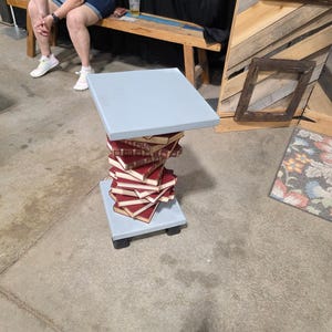 May include: A unique side table made from stacked books. The base and top are light blue, with a stack of red-covered books in between. The table is on a concrete floor.