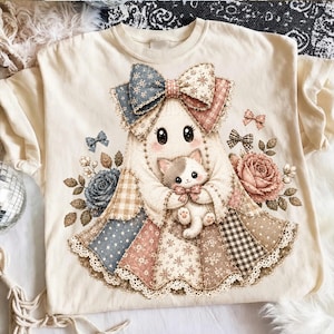 May include: A cream-colored t-shirt featuring a central graphic of a ghost figure wearing a patchwork dress and a large bow. The ghost holds a small kitten. The design includes floral patterns, gingham checks, polka dots, and lace trim details.