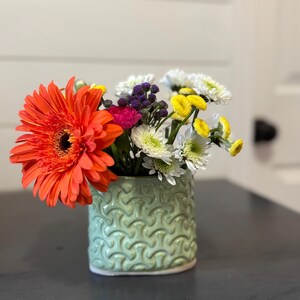 May include: A light green ceramic vase with a repeating embossed geometric pattern holds a vibrant floral arrangement. The bouquet features a large orange gerbera daisy, small white chrysanthemums, yellow button flowers, and purple filler.
