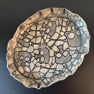 May include: An oval-shaped metal tray with a scalloped edge, featuring a dark, intricate lace-like pattern of skulls and crossbones printed across the surface. The tray has a metallic, silver-toned finish with dark shading that highlights the design.