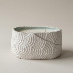 May include: An oval-shaped white ceramic candle vessel with a textured, embossed pattern of overlapping semi-circles. The container features a diagonal seam across the front and holds white wax with two visible cotton wicks centered inside.