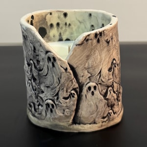 May include: A cylindrical ceramic candle holder with an uneven, open top edge. The exterior surface is off-white with a dark, distressed wash and features a repeating embossed pattern of multiple small ghosts with hollow eyes and mouths.