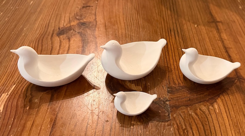 May include: Four white bird-shaped bowls of varying sizes are arranged on a wooden surface. The bowls have a smooth, matte finish and a simple, elegant design. The bowls are ideal for serving snacks or as decorative pieces.