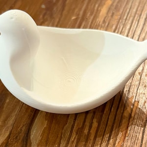 May include: A white bird-shaped dish with a smooth, matte finish. The dish features a bird's head and tail, with a bowl-shaped body. The interior has a textured, concentric circle pattern. The dish is likely made of ceramic or a similar material.
