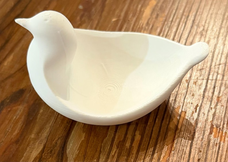 May include: A white bird-shaped dish with a smooth, matte finish. The dish features a bird's head and tail, with a bowl-shaped body. The interior has a textured, concentric circle pattern. The dish is likely made of ceramic or a similar material.