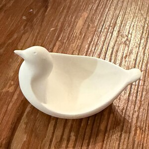 May include: A white ceramic bird-shaped dish, featuring a rounded body and a detailed bird head. The dish is designed with a shallow bowl for holding small items. The bird dish is set against a wooden background.