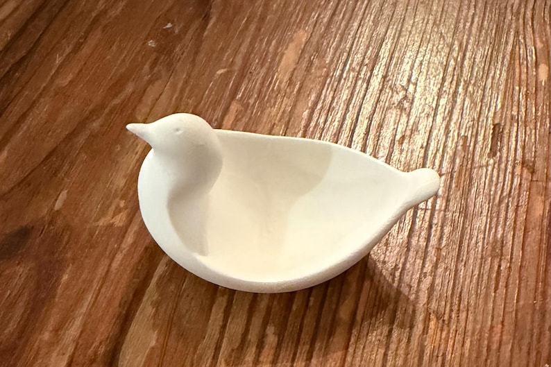 May include: A white ceramic bird-shaped dish, featuring a rounded body and a detailed bird head. The dish is designed with a shallow bowl for holding small items. The bird dish is set against a wooden background.