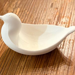 May include: A white, bird-shaped decorative bowl. The bowl has a smooth, curved design with a detailed head and tail. The bowl is sitting on a wooden surface.