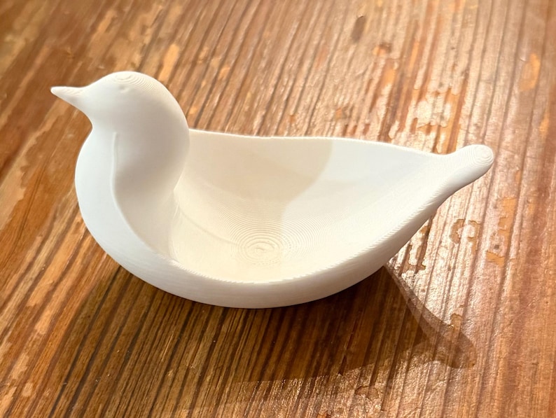 May include: A white, bird-shaped decorative bowl. The bowl has a smooth, curved design with a detailed head and tail. The bowl is sitting on a wooden surface.