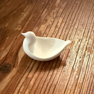 May include: A small, white ceramic bird-shaped dish. The bird has a rounded body, a pointed beak, and a shallow bowl-like interior. The dish is resting on a wooden surface.