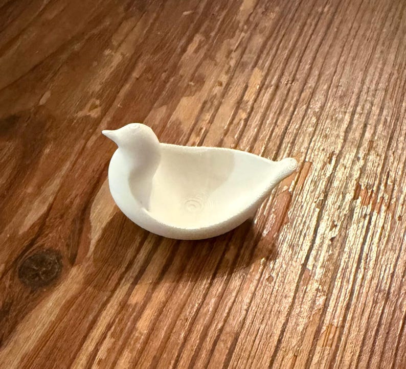 May include: A small, white ceramic bird-shaped dish. The bird has a rounded body, a pointed beak, and a shallow bowl-like interior. The dish is resting on a wooden surface.