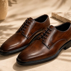 May include: A pair of dark brown leather oxford shoes featuring a burnished finish, a rounded toe, thin dark brown laces, and a solid black sole. The shoes have a smooth, polished surface with visible stitching along the eyelet panels and heel.