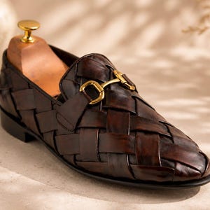 Pode incluir: Um mocassim de couro trançado castanho escuro com um detalhe de bridão em metal dourado polido no peito do pé. O sapato é construído com tiras largas de couro entrelaçadas e inclui uma forma de madeira inserida na área do calcanhar.