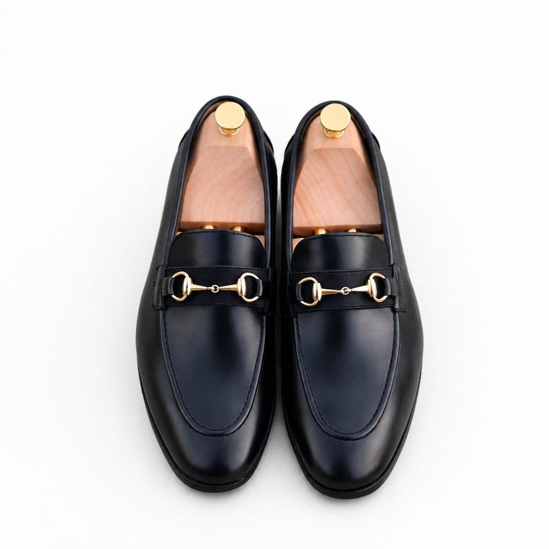 Puede incluir: Un par de mocasines de cuero negro con un detalle de bocado de metal dorado en el empeine. Cada zapato contiene una horma de madera clara con un pomo dorado en el tal&oacute;n, mostrando un acabado liso y un dise&ntilde;o de punta redondeada.