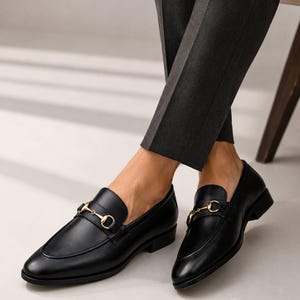 Puede incluir: Un par de mocasines de cuero negro con un detalle de bocado de metal dorado en el empeine. Los zapatos tienen un acabado liso, punta redondeada y tac&oacute;n bajo, combinados con pantalones de color gris oscuro que terminan sobre el tobillo.