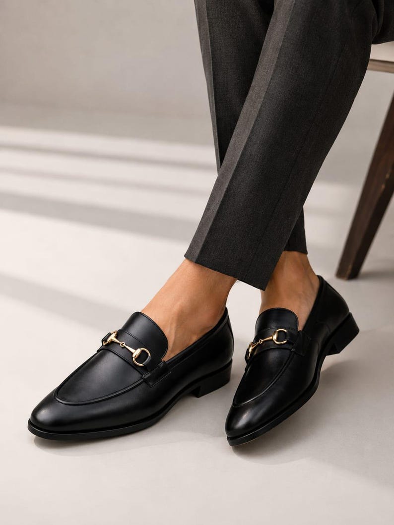 Puede incluir: Un par de mocasines de cuero negro con un detalle de bocado de metal dorado en el empeine. Los zapatos tienen un acabado liso, punta redondeada y tac&oacute;n bajo, combinados con pantalones de color gris oscuro que terminan sobre el tobillo.