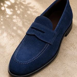 Puede incluir: Un mocasín penny de ante azul marino con una puntera clásica cosida al estilo mocasín, una tira horizontal sobre el empeine y una suela negra sólida. El zapato presenta una textura mate suave y líneas limpias.