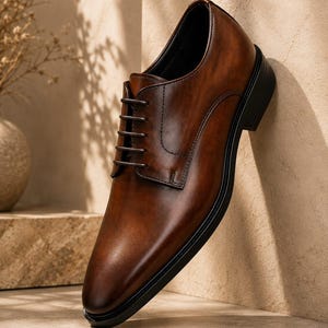 May include: A polished brown leather oxford shoe featuring a smooth, burnished finish, a classic lace-up closure with dark brown laces, a rounded toe, and a solid black sole. The shoe is constructed with visible stitching along the side panels.