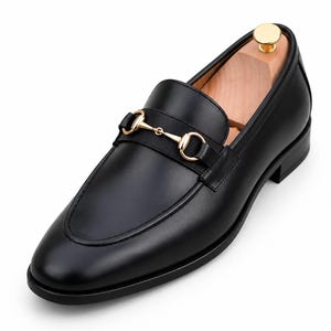 Puede incluir: Un mocas&iacute;n de cuero negro con un detalle de bocado de metal en tono dorado sobre el empeine. El zapato tiene un acabado liso, punta redondeada, tac&oacute;n bajo y una horma de madera clara insertada en el interior para mantener su forma.