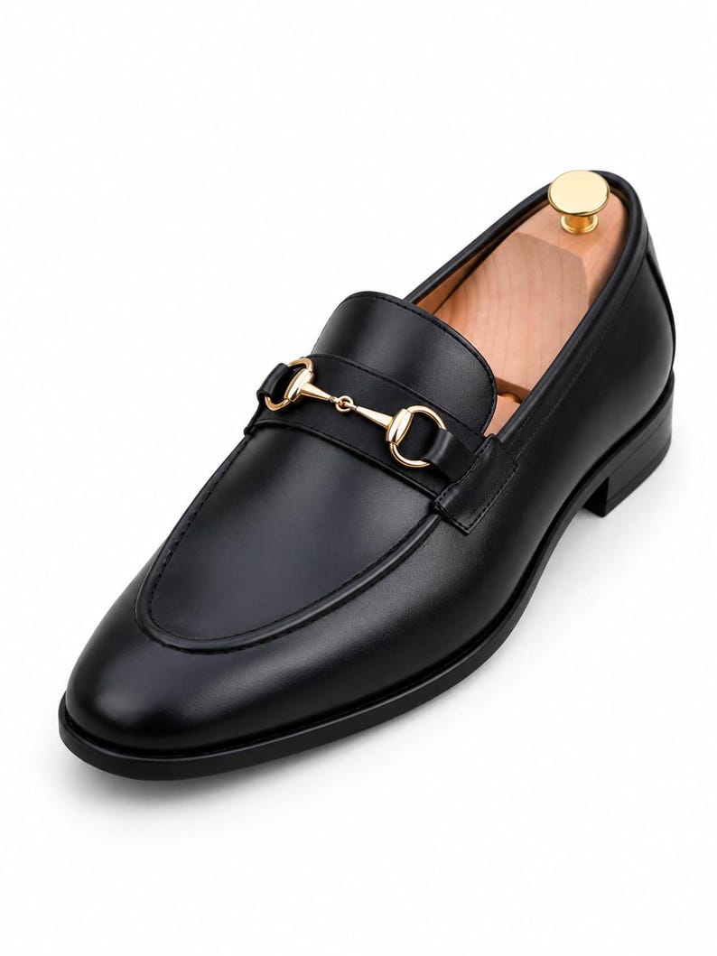 Puede incluir: Un mocas&iacute;n de cuero negro con un detalle de bocado de metal en tono dorado sobre el empeine. El zapato tiene un acabado liso, punta redondeada, tac&oacute;n bajo y una horma de madera clara insertada en el interior para mantener su forma.