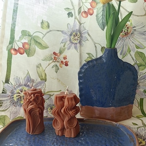Può includere: Due candele marroni scolpite con un design ondulato sono su un vassoio di ceramica blu. Un vaso blu con base marrone contiene due tulipani. Lo sfondo presenta una tenda a motivi floreali con fiori viola e bacche rosse.