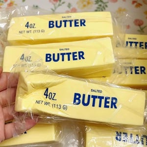 May include: Several rectangular sticks of salted butter wrapped in clear plastic film. Each stick is pale yellow and features blue text that reads 4oz. NET WT. (113 G) SALTED BUTTER. The sticks are stacked together in a group.